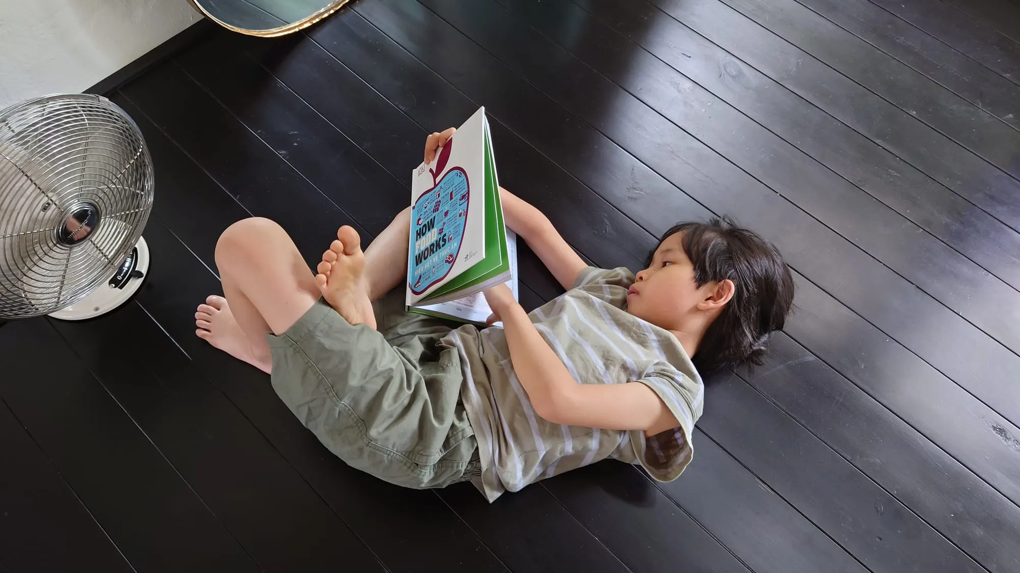 Bé nằm đọc sách trên sàn gỗ